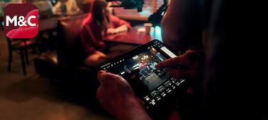 Изображение оператора, который использует планшет для мониторинга камеры FX5, снимающей человека, сидящего за столиком в тускло освещенном кафе.